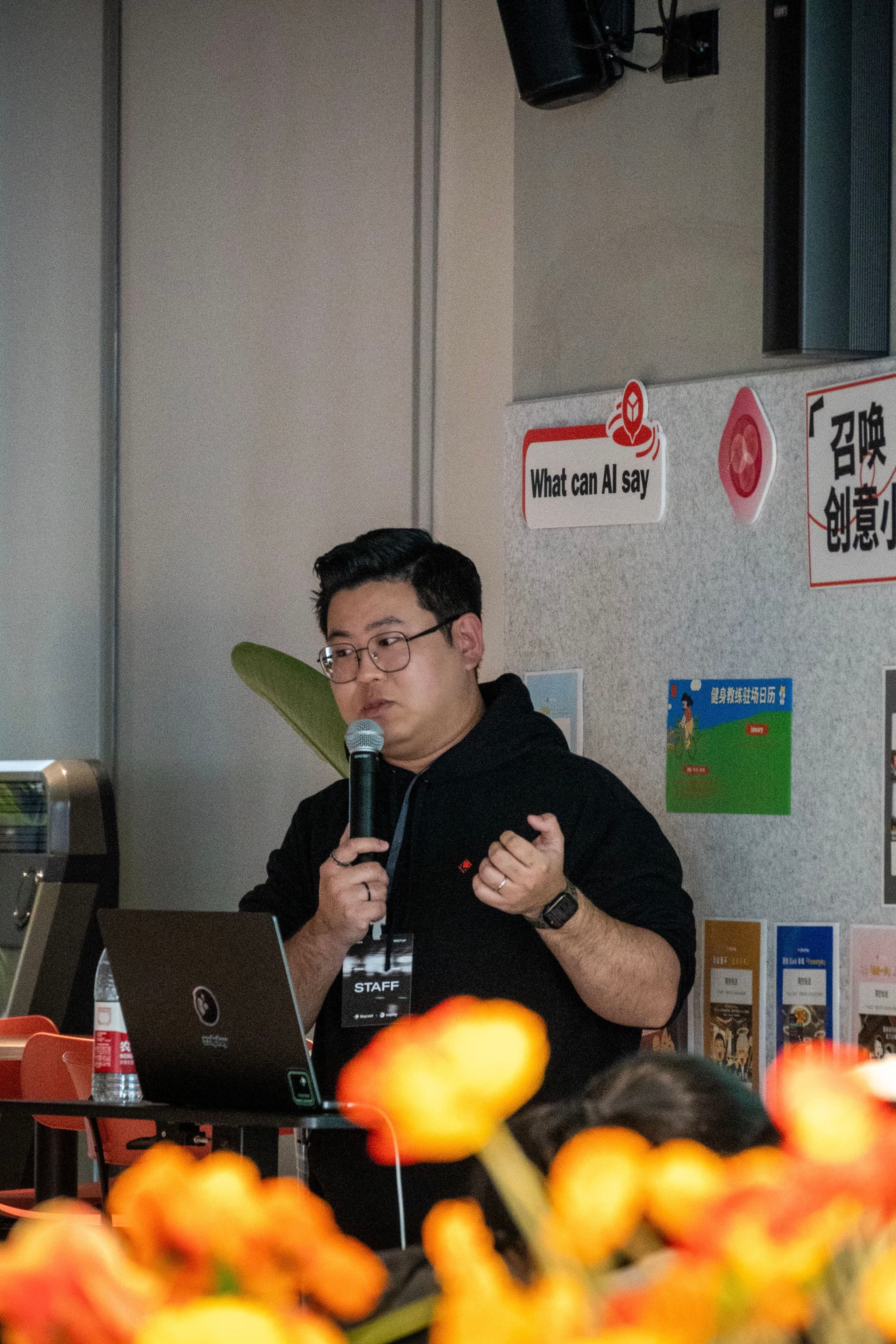 Person holding microphone giving a presentation beside a laptop with posters on the wall and blurred flowers in the foreground