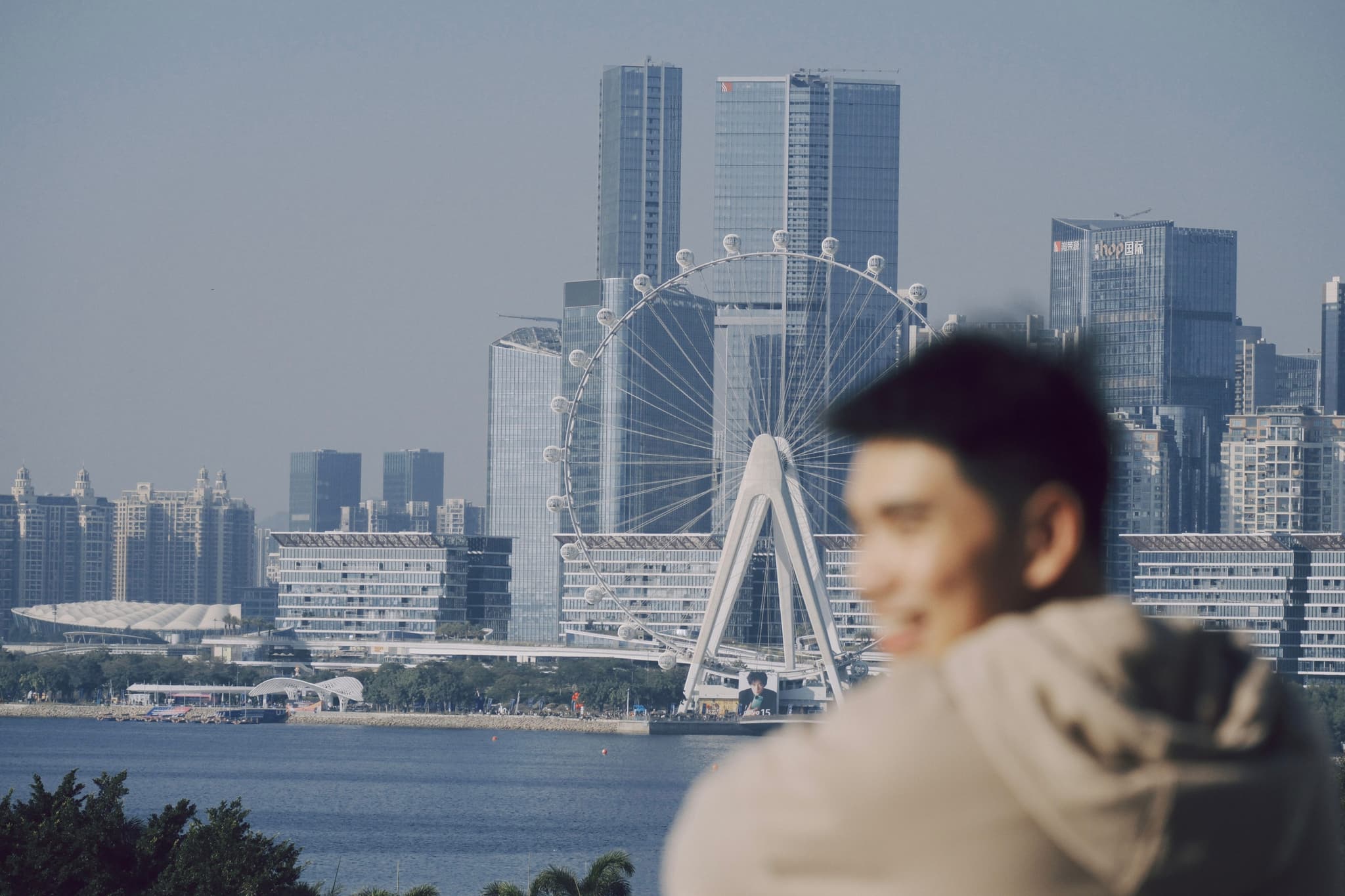 Blurred person in foreground overlooking Qianhai Bay with modern skyline and a distinctive arch bridge in the distance
