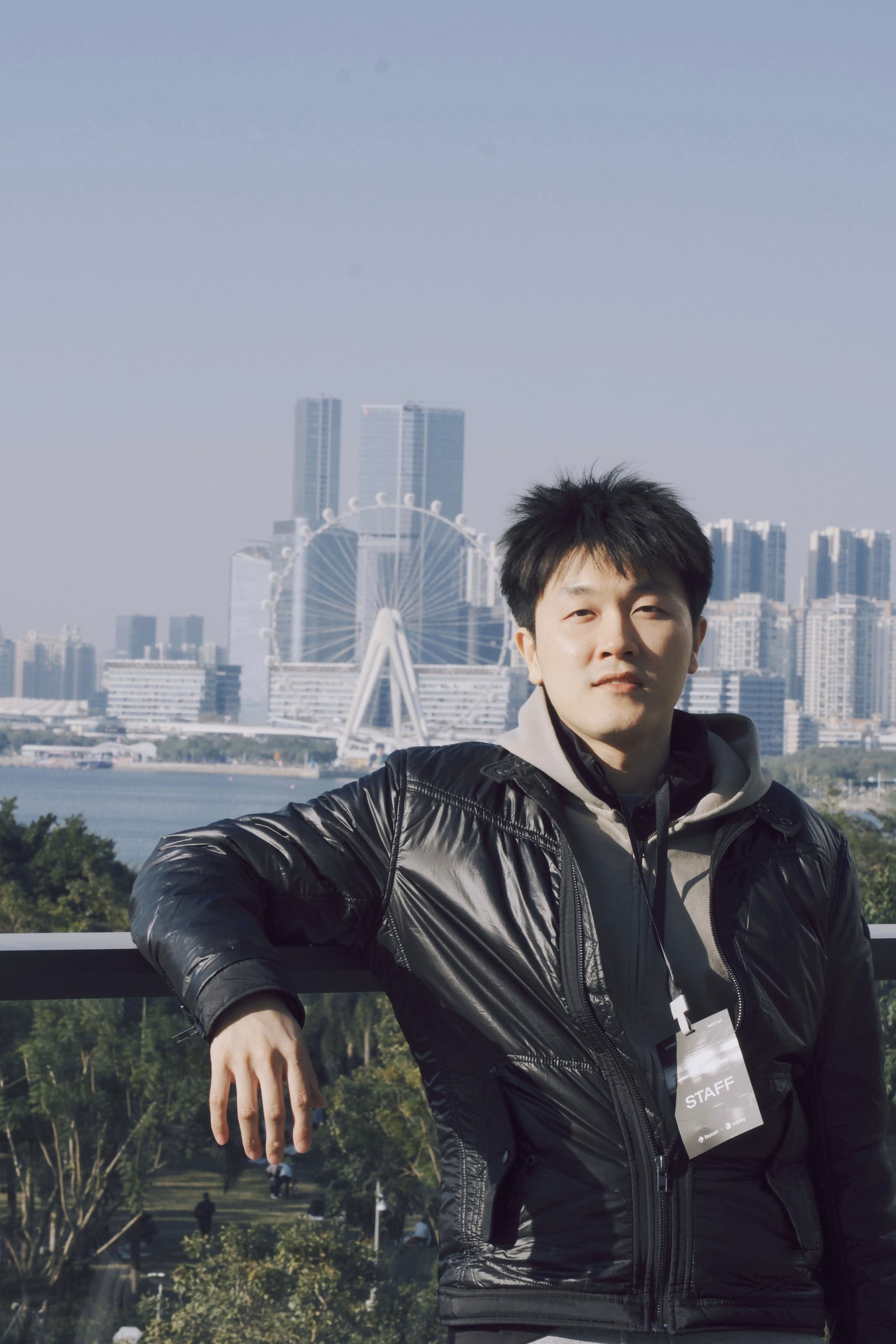 Man in black jacket leaning on balcony railing with city skyline and bridge in the background
